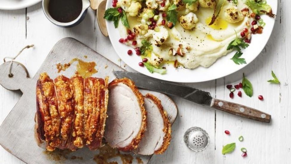 Curtis Stone's Roast Pork on a wooden board with Cauliflower Salad served with mash potato and garnished with pomegranite, cashew nuts, mint and parsley leaves. Gravy to the side.