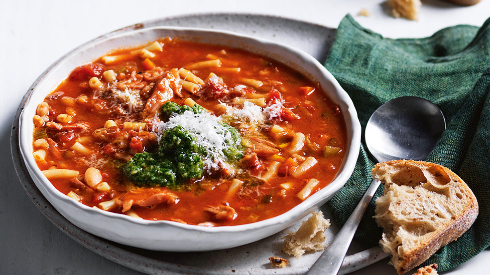 Slow Cooker Ham Hock Minestrone garnished with pesto and cheese, beside bread and green cloth