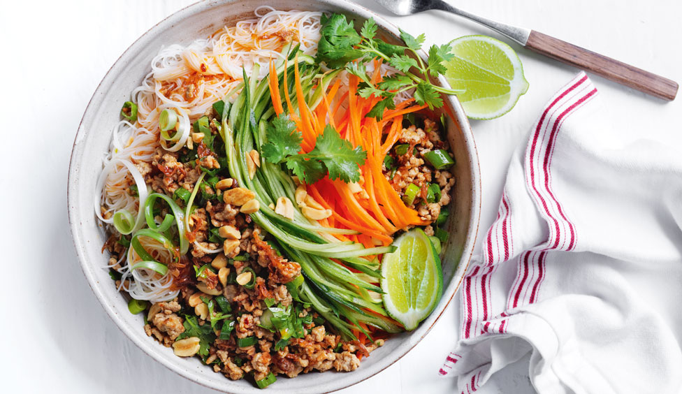 Spicy pork noodle bowl topped with lime wedges, coriander sprigs, carrot and spring onions