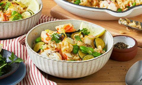 A bowl of lobster risotto with green veggies and lemon wedges garnished with pepper and herbs.