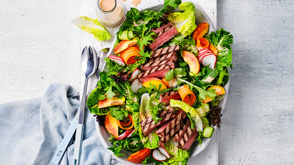 Curtis Stone's BBQ steak salad with Thai vinaigrette on a serving dish, dressed with sliced peaches, shaved carrot and shaved cucumber.