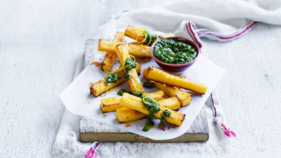 Polenta chips laid out on a serving board with pesto dipping sauce drizzled on top and in a small dish on the side.