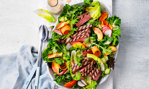 Curtis Stone's BBQ steak salad with Thai vinaigrette on a serving dish, dressed with sliced peaches, shaved carrot and shaved cucumber.