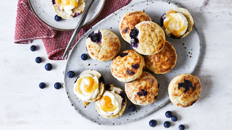 Plate of blueberry and white choclate scones with blueberries scattered across the kitchen bench