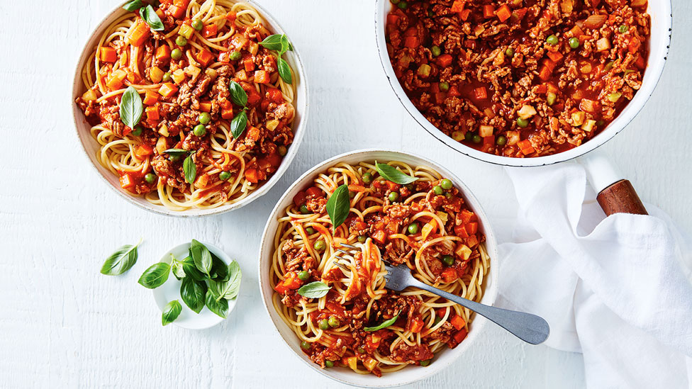 Two bowls and a pot of veggie loaded spaghetti bolognese with basil on the side