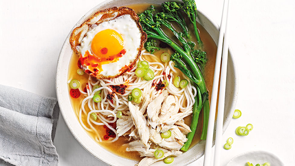 A bowl of soup with noodles, chicken, greens and a fried egg