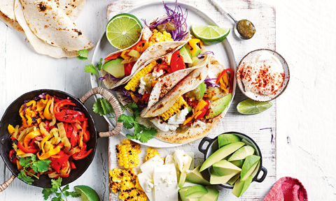 Three tacos served on a plate with corn, avocado, lime wedges and chillies on the side