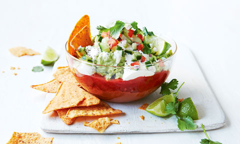 Mexican dip in a bowl topped with corn chips, cucumber, tomato, coriander and lime wedges