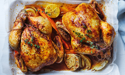 Two roast chickens in a baking tray with lemon, carrot, garlic and thyme