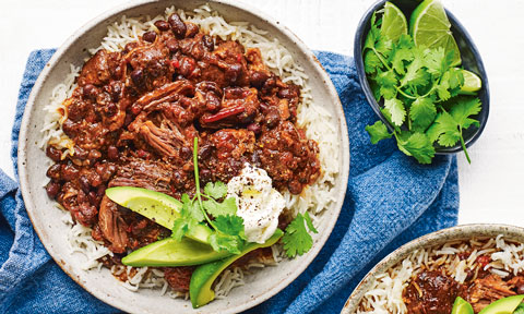 Beef and black bean chilli served on a bed of rice with avocado garnish and lime wedges and coriander on the side