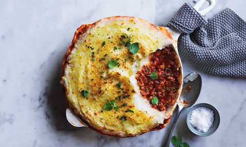 Curtis Stone's Nonna's Cottage Pie garnished with oregano leaves in an oven dish. Salt to the side, arranged on a marble table with a grey towel. 