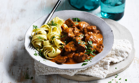 Slow-cooker Beef Stroganoff in dish sprinkled with parsley. On a wooden board with white cloth.