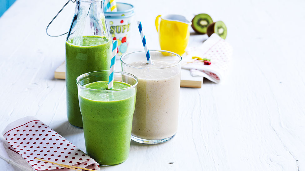 Spinach smoothies in glasses, with a jug of smoothie and chopped fruit on side