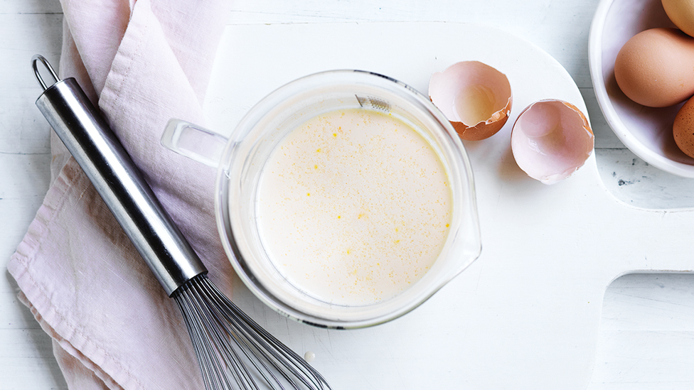 A bowl of pancake mixture with an whisk and cracked eggs on the side