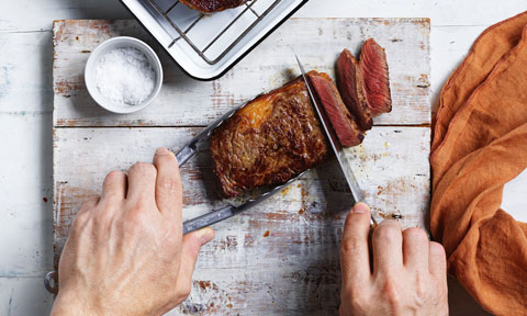 Cooked steak being sliced on a wooden chopping board with salt on the side