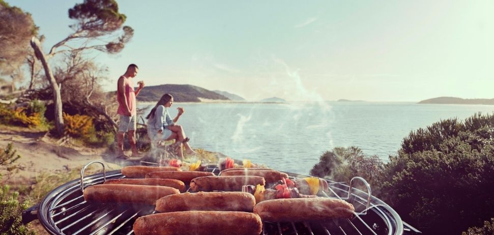 Sausages on a BBQ with two people at the beach in the background